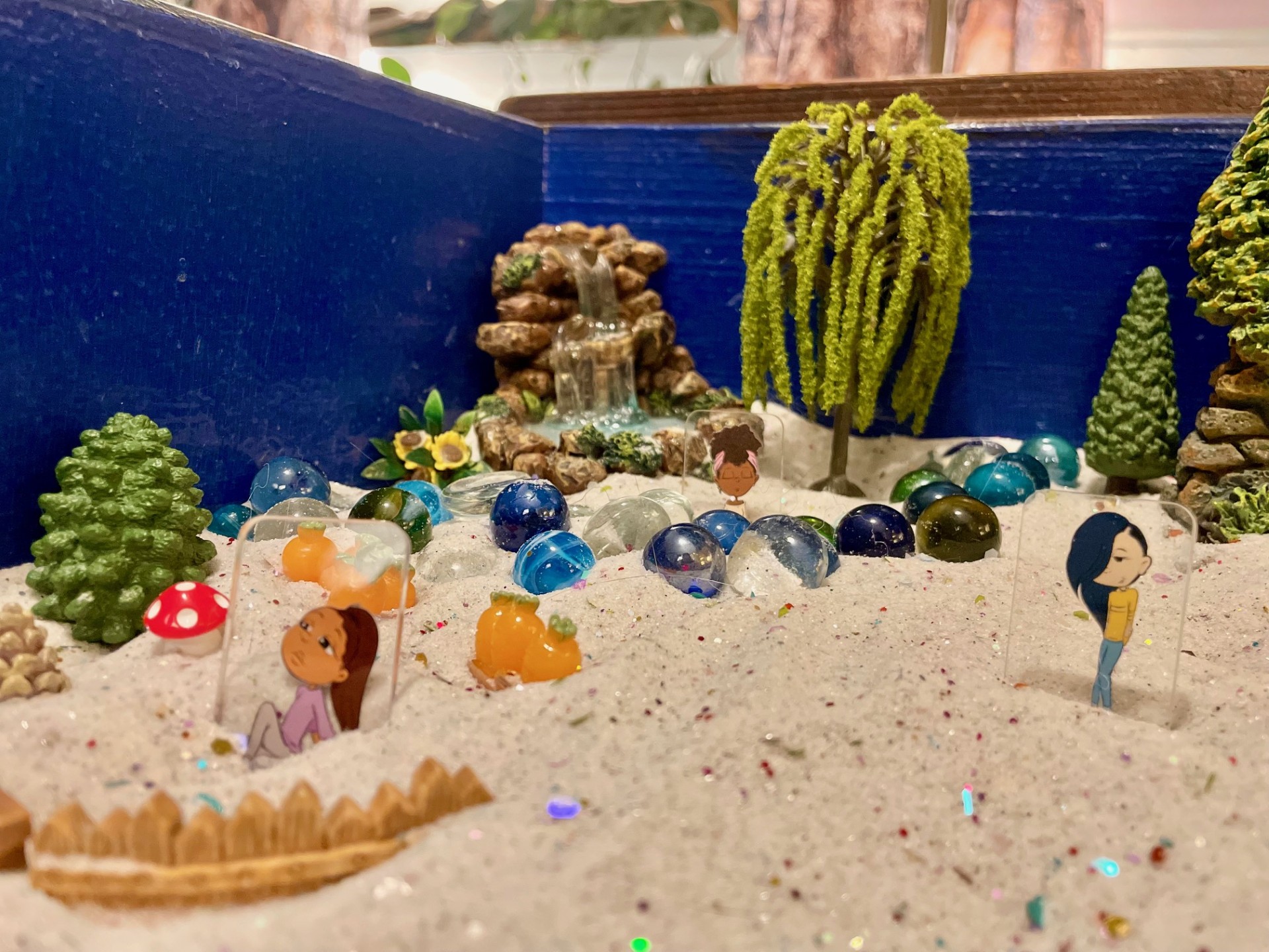 Sand tray with people figurines with different facial expressions (relaxed, pondering, and chill) with trees in the background, a waterfall, a river, and a garden with carrots, a mushroom, and acorn.