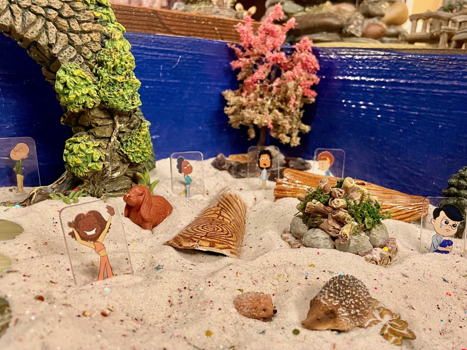 Sand tray with people figurines with different facial expressions (happy, disgruntled, confused, sad, and wondering) with trees in the background, and logs around an unlit campfire. There are also two hedgehogs and a bunny.